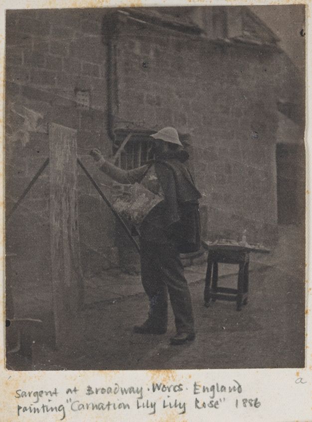 Black and white photograph. A man stands at an easel, painting, with a palette resting along the other arm. He is in front of a stone building. A pencil notation under the image reads, “Sergeant at Broadway Worcs England Painting ‘Carnation Lily Lily Rose’ 1886”.