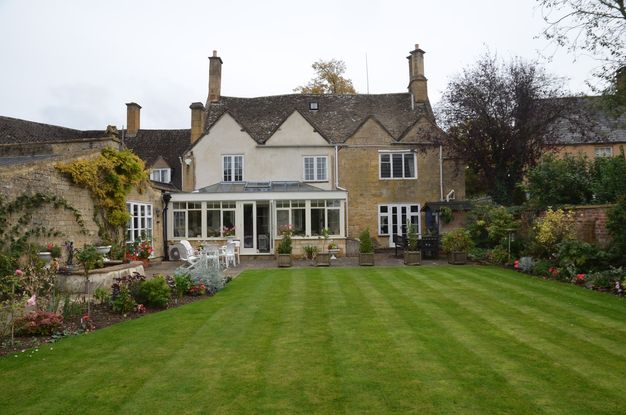 Colour photograph. A four-peaked house is flanked by gardens to either side of a tidily mowed green lawn. The left two bays of the house are cream white and have a sunroom projecting from the back. The other two bays are brown stone.